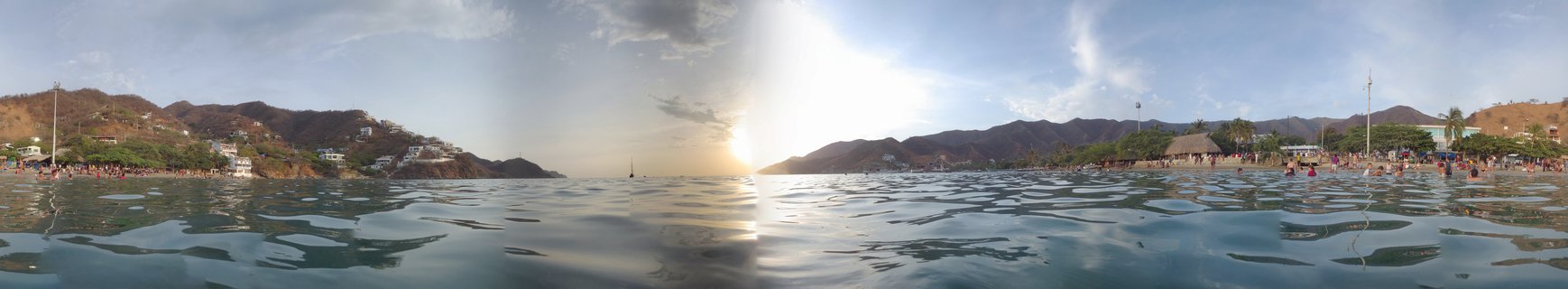 Panoramic beach scene at sunset showing calm Caribbean water in foreground, people along shoreline, white buildings on brown mountainside, and sun setting between mountain peaks