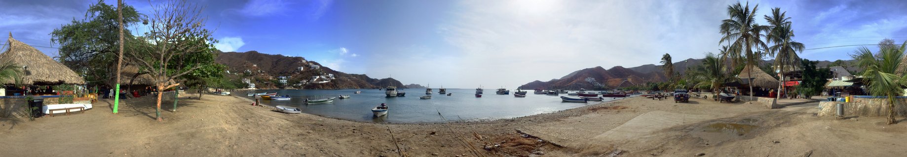 Panoramic view of a tropical bay with multiple fishing boats anchored in calm blue water, sandy beach foreground with palm trees and thatched-roof huts, hills in background under partly cloudy sky