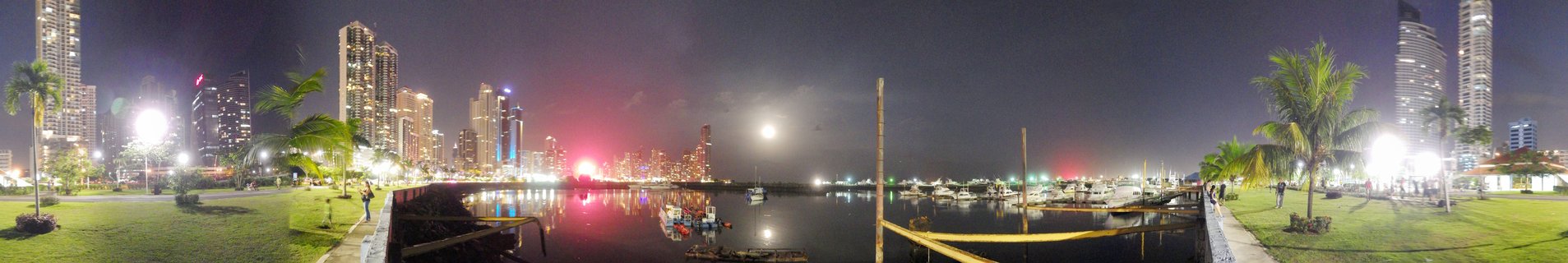 Night panoramic view of Panama City skyline with illuminated high-rise buildings, marina with boats, palm trees, and waterfront park with people walking along pathways