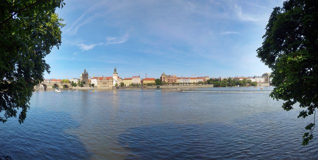 Panoramic view of Charles Bridge spanning Vltava River in Prague with historic Old Town buildings, Gothic tower, and blue sky with wispy clouds