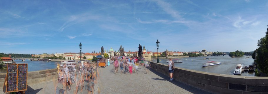 Tourists walk across Prague's historic Charles Bridge spanning the Vltava River, with baroque statues, street vendors, and the city's red-roofed Old Town visible under a blue summer sky.