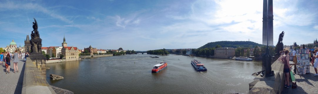 Panoramic view from Charles Bridge showing the Vltava River with two tour boats, baroque statues on the bridge, historic buildings with red roofs, and Prague's Old Town skyline under a blue summer sky