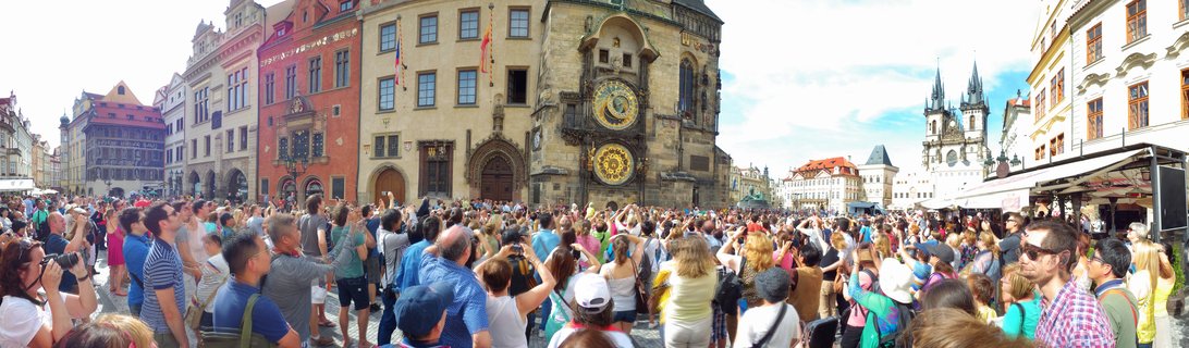 Large crowd of tourists in historic European square looking at ornate astronomical clock on medieval stone tower with Gothic church spires in background