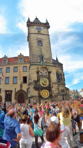 A tall medieval stone clock tower with two ornate golden astronomical clock faces, surrounded by a large crowd of tourists on a cobblestone square under a blue sky with clouds