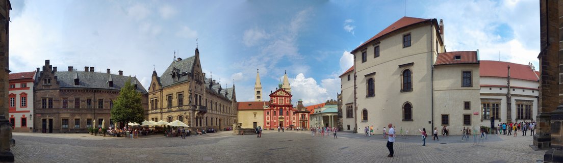 Wide view of Prague Castle Third Courtyard showing historic buildings with red roofs, cobblestone ground, tourists walking, and blue sky with clouds