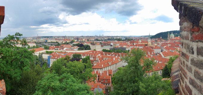 Panoramic cityscape of Prague showing red-tiled roofs, historic buildings, church towers, and green trees under cloudy sky