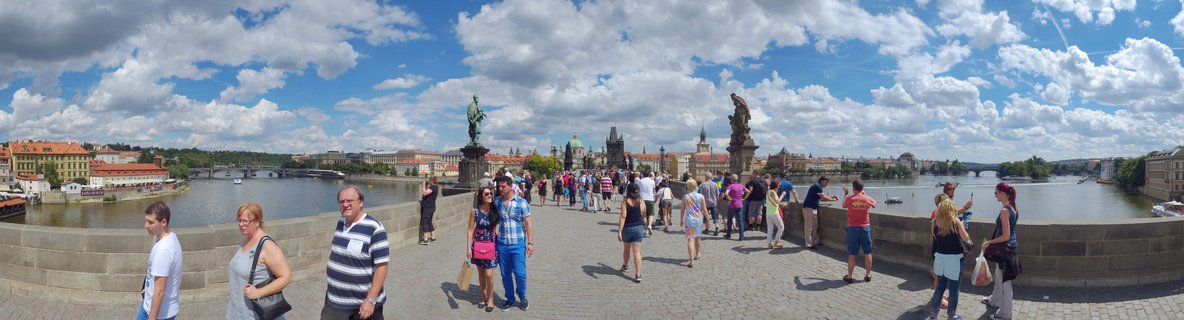 Panoramic view of tourists walking on the historic stone Charles Bridge in Prague with baroque statues, the Vltava River, and red-roofed buildings under a cloudy blue sky