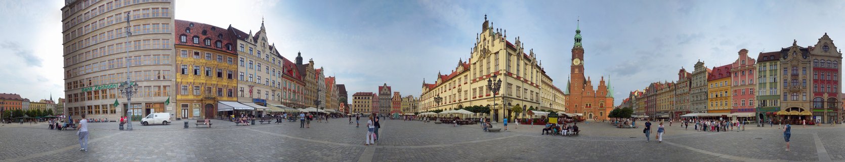 Wide panoramic photograph of Wrocław's Old Town Market Square showing the Gothic Town Hall with tall clock tower, rows of colorful historic buildings with red tile roofs, cobblestone plaza with scattered pedestrians under partly cloudy sky