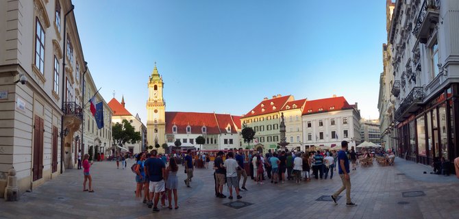 Wide panoramic view of Bratislava's Main Square showing the Old Town Hall tower, historic buildings with red roofs, and numerous tourists walking on the paved plaza under clear blue sky