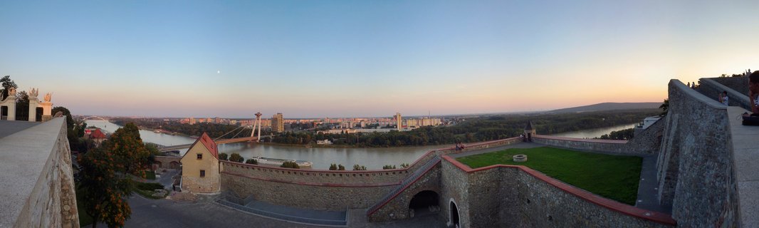 Panoramic cityscape view from Bratislava Castle showing stone fortress walls in foreground, Danube River flowing through center, UFO Bridge tower bridge, and Bratislava city skyline at sunset with gradient sky