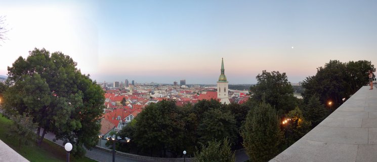 Wide panoramic cityscape of Bratislava at dusk showing St. Martin's Cathedral tower with green spire, red-tiled rooftops, foreground trees, and twilight sky with visible moon