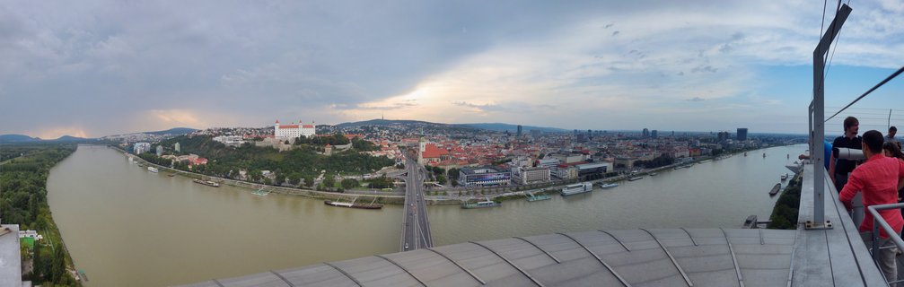 Wide panoramic photograph showing Bratislava Castle on a green hill overlooking the brown-green Danube River with city buildings, bridges, and boats under a cloudy sky with sun rays breaking through