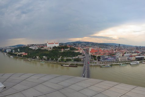 Panoramic cityscape showing Bratislava Castle on a green hill above the Danube River with red-roofed Old Town buildings, a bridge crossing the water, and river cruise ships docked along the shore under cloudy skies