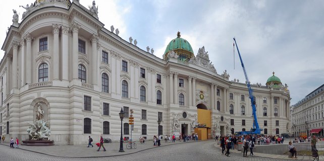 Wide view of the Hofburg Palace in Vienna showing the white baroque building with green domes, ornate sculptures, and tourists walking on cobblestone courtyard under cloudy sky