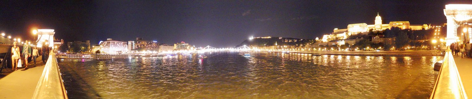 Panoramic night photograph of Budapest showing the illuminated Buda Castle on a hill overlooking the Danube River, with the Chain Bridge's stone towers and suspension cables visible, city lights reflecting on the water