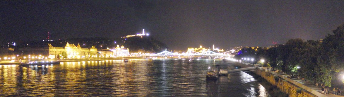 Night panoramic view of Budapest showing the illuminated Chain Bridge spanning the Danube River with Buda Castle and Gellért Hill visible on the horizon, golden building lights reflecting on water