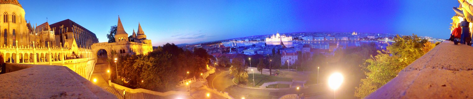 Panoramic night view of Budapest showing illuminated Fisherman's Bastion stone towers on the left and Hungarian Parliament Building across the Danube River under deep blue twilight sky