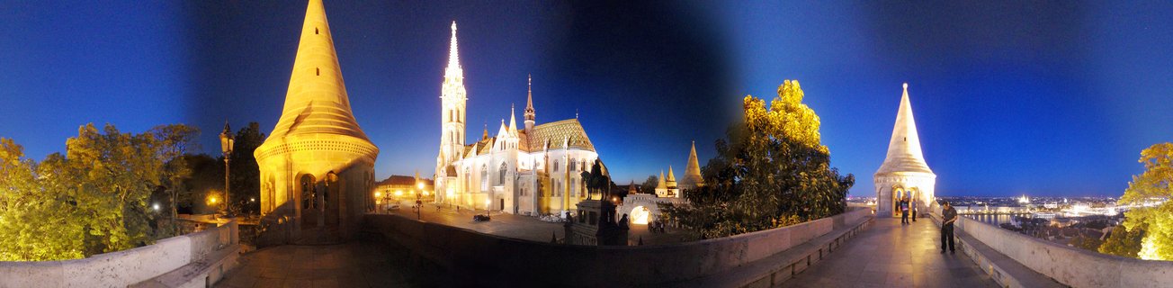 Panoramic view of illuminated Fisherman's Bastion towers and Matthias Church at blue hour in Budapest with tourists on stone walkways and city lights visible in the distance