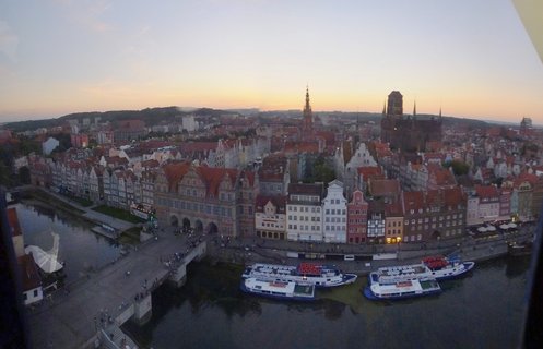 Elevated panoramic view of Gdańsk Old Town showing colorful historic buildings along the Motława River with tourist boats docked at the waterfront and church spires visible against a sunset sky