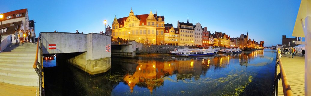Panoramic view of Gdansk Old Town buildings along Motława River at blue hour with illuminated historic architecture, boats, and reflections in water