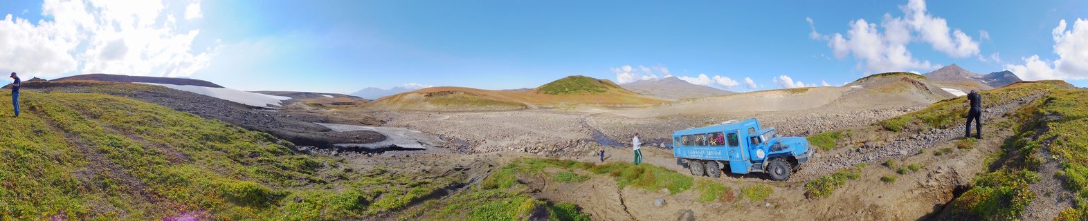 Blue all-terrain expedition vehicle on rocky tundra landscape in Kamchatka, Russia with mountains, snow patches, and photographers in the scene