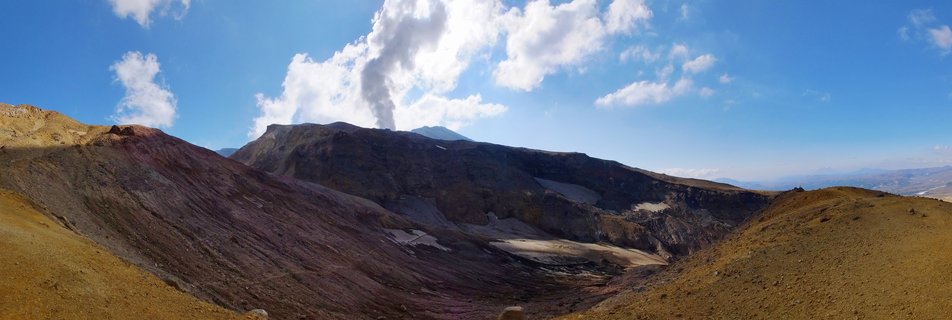 Volcanic crater landscape with steam plume rising from mountain ridge against blue sky with white clouds in Kamchatka Russia