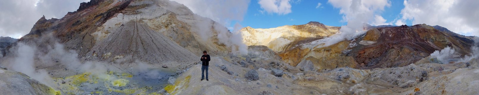 Person standing in geothermal valley with steam vents rising from colorful yellow, red and white mineral-stained volcanic mountains under blue sky with clouds