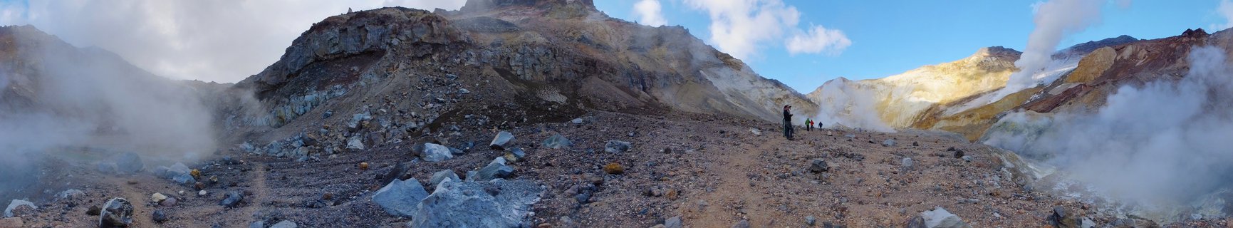 Panoramic view of geothermal fumaroles releasing white steam across rocky volcanic terrain with colorful mineral deposits in Kamchatka, Russia