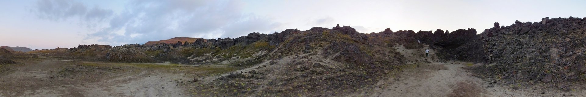 Panoramic view of a volcanic landscape in Kamchatka, Russia, showing dark lava rock formations, patches of green moss and lichen, a dirt trail winding through the terrain, and a lone hiker walking on the right side under an overcast sky