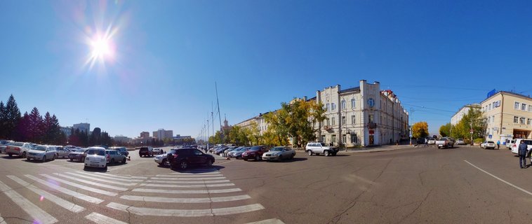 Wide street scene in Ulan-Ude, Russia showing parked cars, crosswalk, multi-story buildings, trees with autumn foliage, and bright sun in clear blue sky
