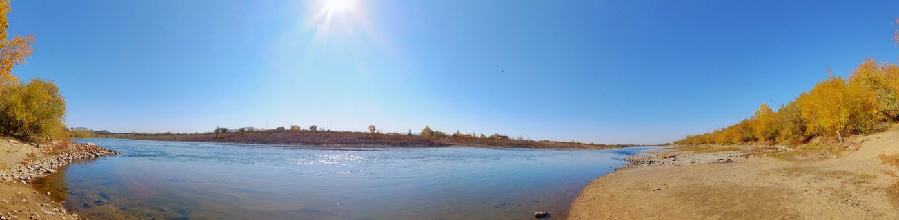 Wide panoramic photograph of a river with golden autumn trees on both banks under a bright blue sky with the sun visible overhead