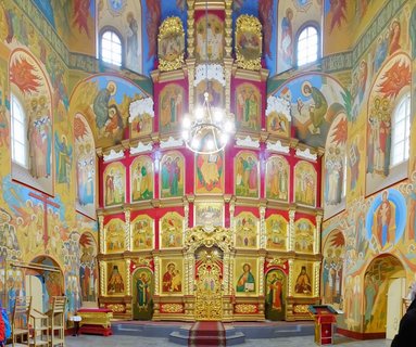 Interior of Russian Orthodox church showing ornate golden iconostasis with multiple rows of religious icons, colorful wall frescoes of saints, arched windows, and hanging chandelier