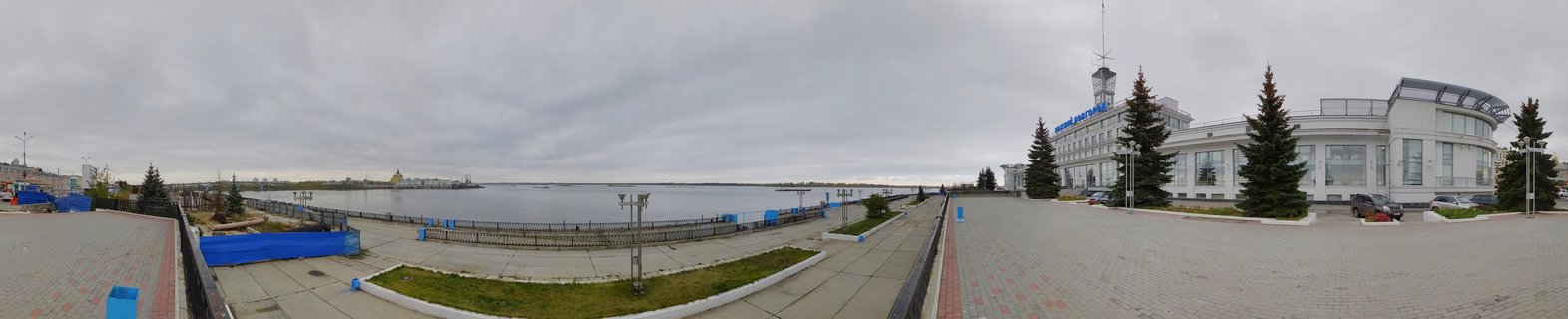 Wide panoramic photograph of Nizhny Novgorod River Terminal building with white facade, paved embankment promenade, evergreen trees, and Volga River under overcast sky