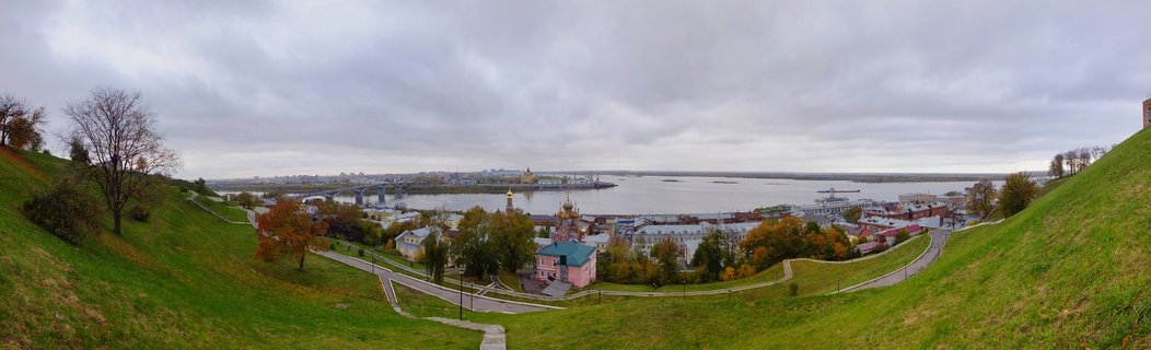 Panoramic landscape showing Nizhny Novgorod Russia with green grassy hills in foreground, golden-domed Orthodox church, city buildings, wide Volga River, and overcast autumn sky