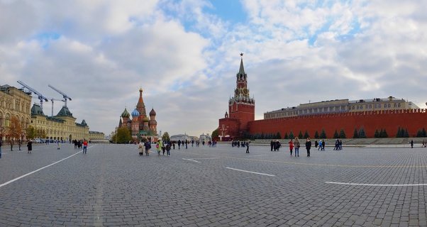 Wide view of Red Square in Moscow showing Saint Basil's Cathedral with colorful domes, the Kremlin's red brick walls and Spasskaya Tower with clock, cobblestone plaza with scattered visitors, and cloudy sky