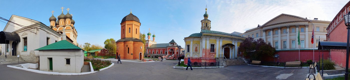 Panoramic courtyard view of Moscow Kremlin showing Russian Orthodox churches with onion domes, red brick towers, and historic buildings with people walking on paved ground under blue sky