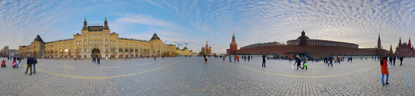 Panoramic photograph of Red Square in Moscow showing GUM Department Store, Kremlin walls, and Saint Basil's Cathedral with pedestrians on cobblestone plaza under cloudy sky