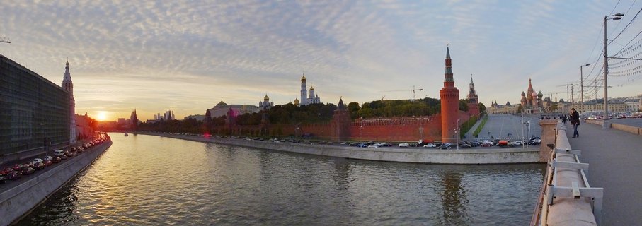 Panoramic view of Moscow Kremlin red brick walls and towers with Saint Basil's Cathedral colorful onion domes at sunset, Moskva River in foreground with reflections, bridge with pedestrians on right side