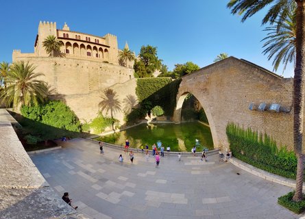 Historic stone palace with crenellated walls, arched bridge over green reflecting pool, palm trees, and tourists on paved plaza under blue sky