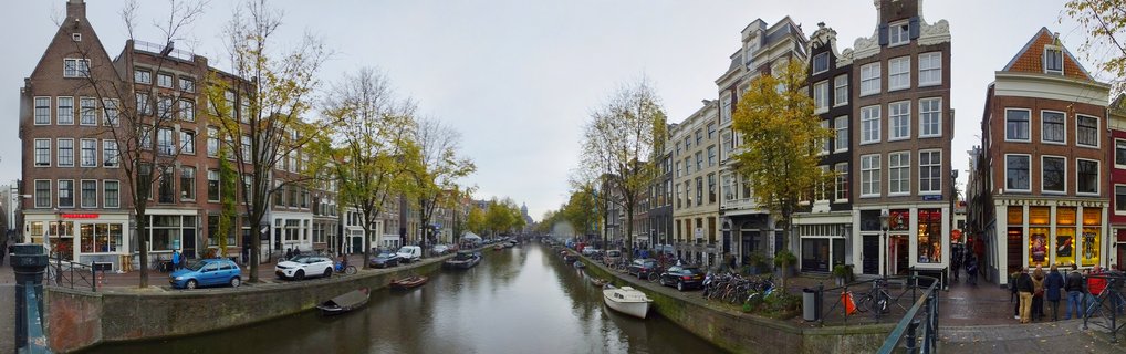 Panoramic view of Amsterdam canal with traditional Dutch gabled houses on both sides, autumn trees with yellow leaves, boats in the water, bicycles and cars parked along the streets, people walking on sidewalks