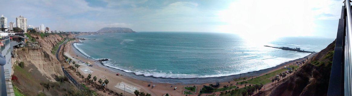 Panoramic aerial view of Lima's coastline showing cliffs with buildings, a sandy beach, palm trees, roads, and the Pacific Ocean with waves under a bright sky