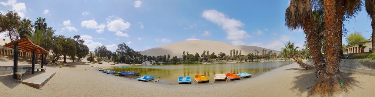 Panoramic view of Huacachina oasis in Peru showing a green lagoon with colorful paddle boats, palm trees, buildings, and large sand dunes under a blue sky with white clouds