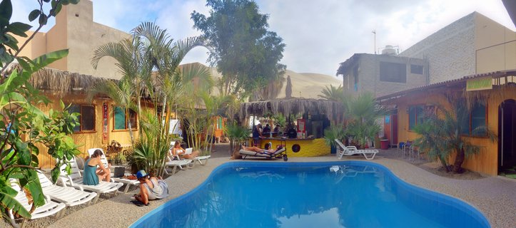 Swimming pool at a resort in Huacachina desert oasis with people lounging on white chairs, palm trees, thatched-roof buildings, and large sand dunes in the background