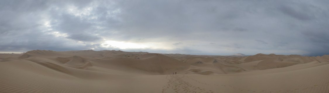 Panoramic view of rolling sand dunes in Huacachina desert with overcast cloudy sky and two small human figures walking in the distance