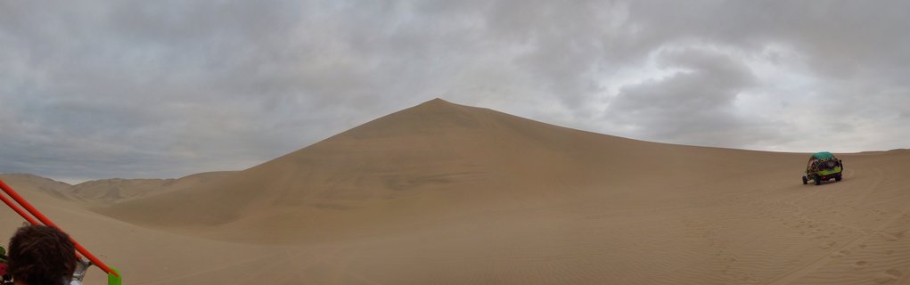 Large sand dune under cloudy sky with two dune buggies carrying tourists across the desert landscape in Huacachina, Peru