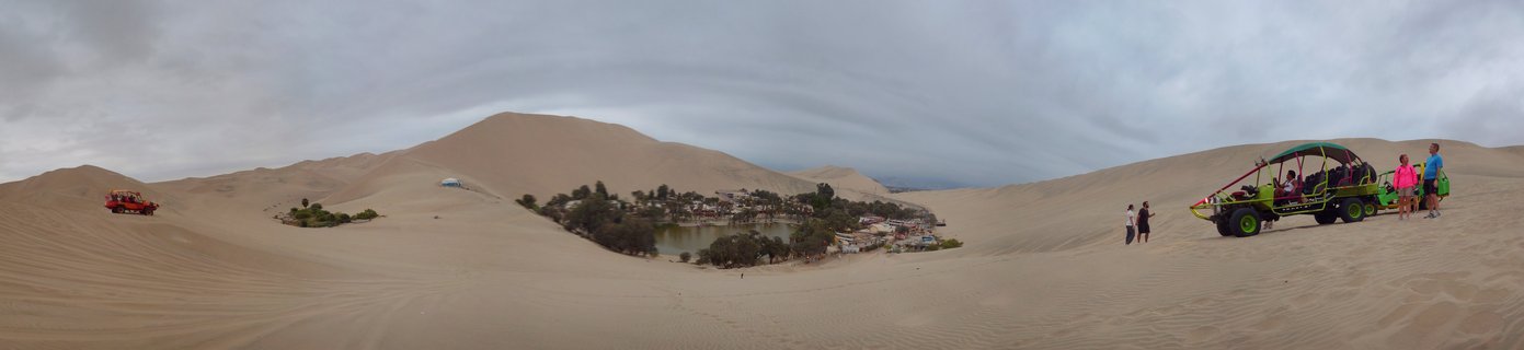 Wide panoramic desert landscape showing Huacachina oasis village with green lagoon and palm trees surrounded by large sand dunes, with colorful dune buggies and tourists visible on the sand under cloudy sky
