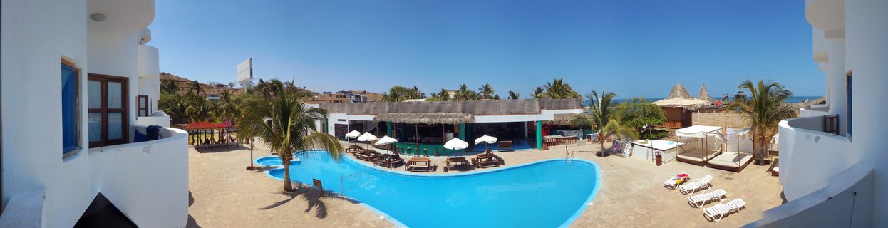 Resort swimming pool with blue water surrounded by white lounge chairs, palm trees, and white buildings with ocean visible in background under clear blue sky