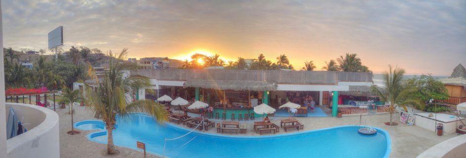Panoramic view of a beach resort with a large blue swimming pool, thatched-roof bar building, palm trees, and sunset over the ocean in Mancora, Peru