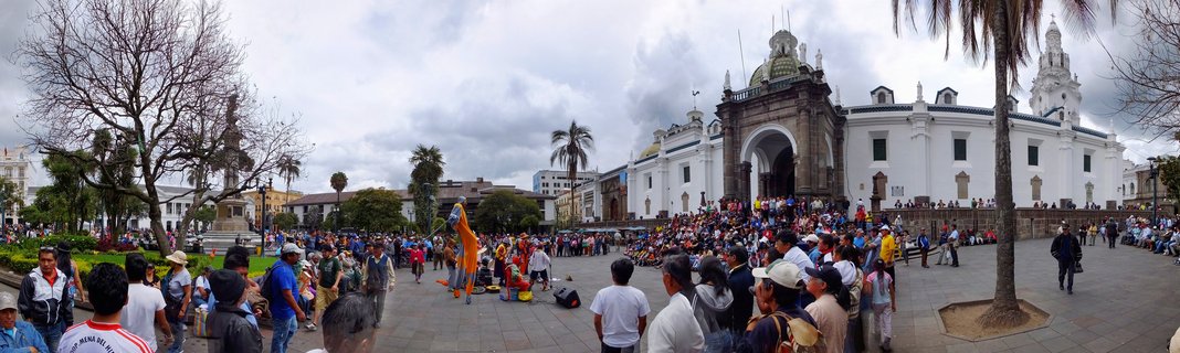 Wide view of a crowded public plaza with a white cathedral featuring a green dome, people gathered throughout the square, street performers in colorful costumes, palm trees, and colonial buildings under an overcast sky