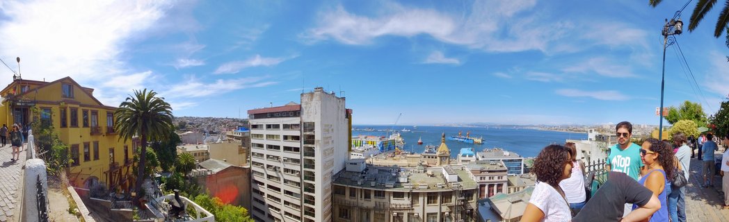 Panoramic view of Valparaíso Chile showing colorful hillside buildings descending toward Pacific Ocean harbor with cargo ships, palm trees, and tourists at viewpoint under blue sky with wispy clouds